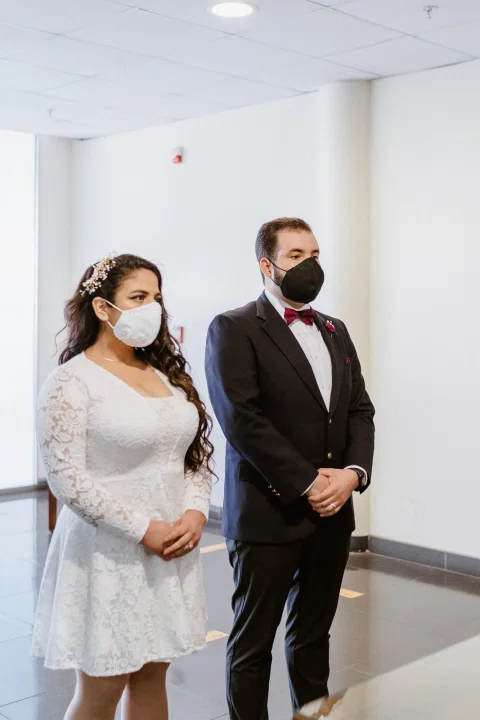 Mujer con vestido blanco y hombre con traje oscuro permanecen de pie juntos dentro de una sala clara.