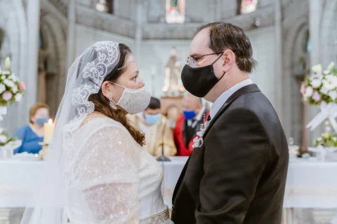 Mujer con vestido blanco y hombre con traje oscuro se miran de cerca mientras usan mascarillas.