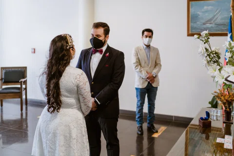 Mujer con vestido blanco y hombre con traje oscuro permanecen frente a frente dentro de una sala, con otra persona al fondo.