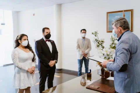 Mujer con vestido blanco y hombre con traje oscuro permanecen frente a un hombre que lee un documento dentro de una sala.