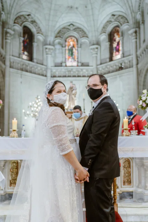 Mujer con vestido blanco y hombre con traje oscuro posan tomados de la mano frente al altar de una iglesia.