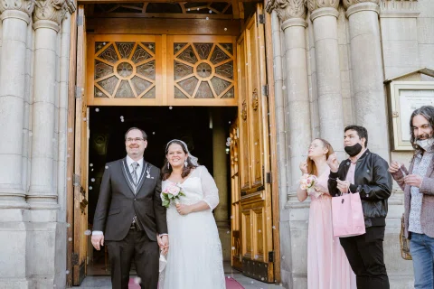 Mujer con vestido blanco y hombre con traje oscuro salen de una iglesia entre varias personas reunidas en la entrada.