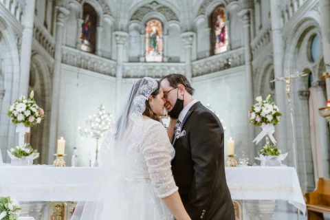 Mujer con vestido blanco y hombre con traje oscuro se besan frente al altar de una iglesia.