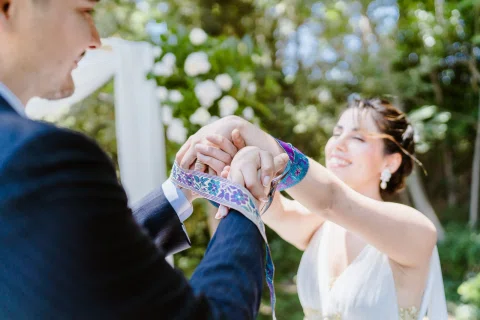 Mujer con vestido blanco y hombre con traje oscuro sostienen una cinta colorida alrededor de sus manos durante la ceremonia.
