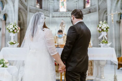 Mujer con vestido blanco y hombre con traje oscuro, ambos con mascarilla, permanecen tomados de las manos frente al altar.