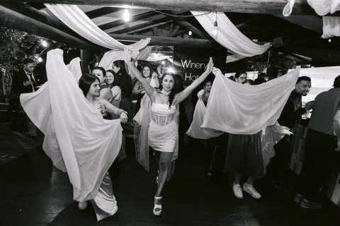 Mujer con vestido claro camina entre dos personas que sostienen telas amplias, en una fotografía en blanco y negro.