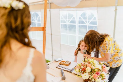 Mujer con vestido floral acerca un micrófono a una niña sentada junto a una mesa con flores y documentos.