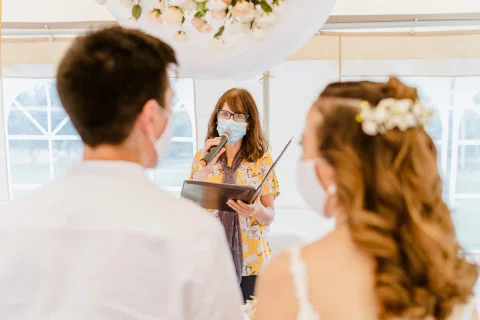 Mujer con vestido floral, mascarilla y lentes habla por un micrófono mientras sostiene una carpeta frente a un hombre y una mujer vistos de espaldas.