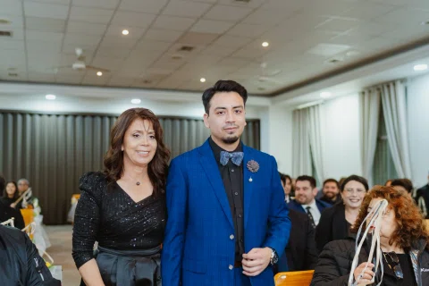 Mujer con vestido negro y hombre con traje azul caminan juntos dentro de una sala con personas sentadas.