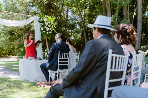 Mujer con vestido rojo habla frente a varias personas sentadas en un jardín.