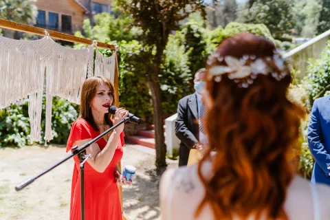 Mujer con vestido rojo habla por un micrófono frente a una mujer con vestido blanco en el jardín.