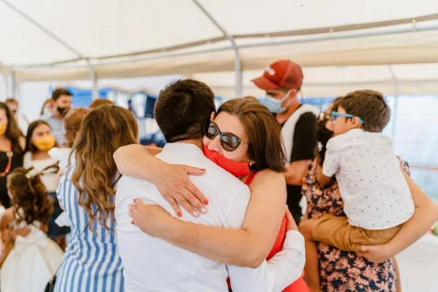 Mujer con vestido rojo y mascarilla abraza a un hombre con camisa blanca; otras personas aparecen alrededor.