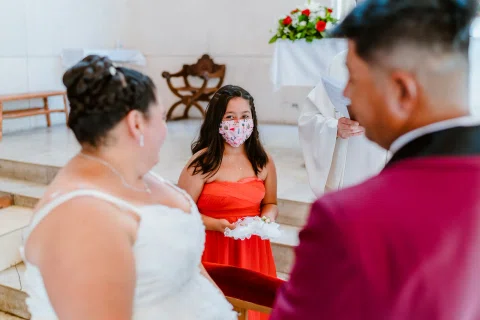 Mujer con vestido rojo y mascarilla frente a una mujer con vestido blanco y un hombre con traje burdeos.