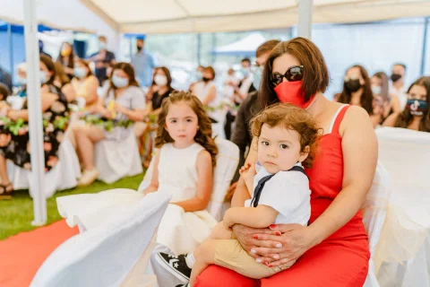 Mujer con vestido rojo y mascarilla está sentada con un niño pequeño en el regazo y una niña a su lado.
