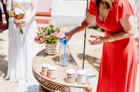 Mujer con vestido rojo vierte material azul desde un recipiente pequeño dentro de un frasco de vidrio sobre una mesa.