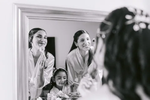 Dos mujeres sonríen y una niña aparece entre ellas en el reflejo de un espejo, con otra mujer desenfocada en primer plano, en blanco y negro.