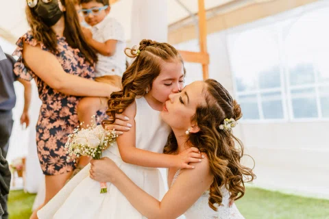 Niña con vestido blanco besa la mejilla de una mujer con vestido blanco que sostiene un ramo.