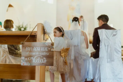 Niña de vestido claro está junto a un cartel de madera al costado de una banca dentro de una iglesia.