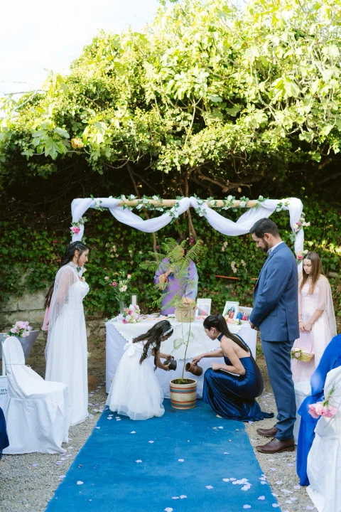 Niña con vestido blanco y mujer con vestido azul vierten contenido desde dos jarras en una maceta con un árbol joven; un hombre con traje azul y una mujer con vestido blanco observan.