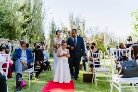 Niña con vestido blanco camina por una alfombra roja junto a un hombre con traje oscuro.