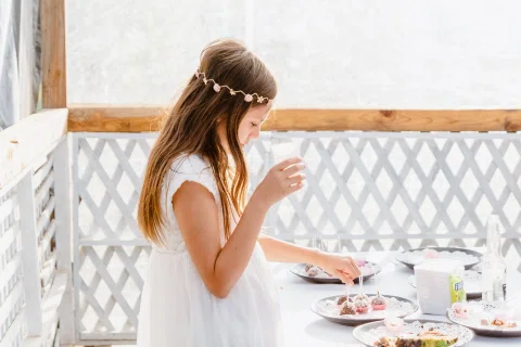 Niña con vestido blanco está junto a una mesa con dulces y sostiene un pequeño recipiente en la mano.
