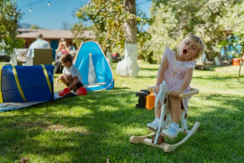 Niña con vestido claro juega sobre un caballo balancín de madera en el césped.