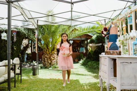 Niña con vestido rosado esparce pétalos desde una canasta mientras camina por el jardín.