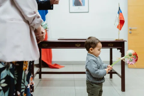 Niño pequeño sostiene flores junto a una mesa, con una bandera de Chile al fondo.