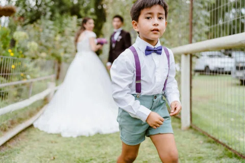 Niño con suspensores y corbatín camina en primer plano, con un hombre y una mujer desenfocados al fondo.