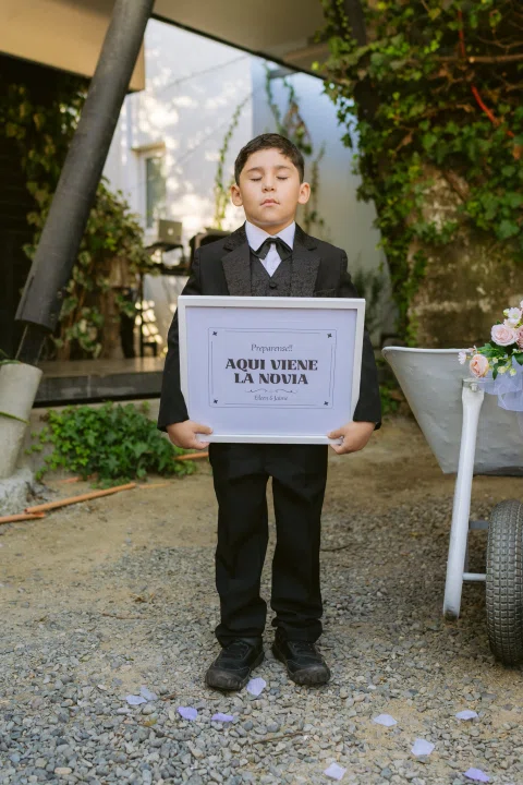 Niño con traje oscuro sostiene un cartel frente a una zona ajardinada.