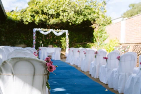 Pasillo con alfombra azul, filas de sillas blancas y un arco floral al fondo.