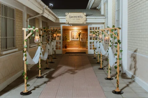 Pasillo exterior de acceso decorado con telas blancas, vegetación y pequeños frascos colgantes.