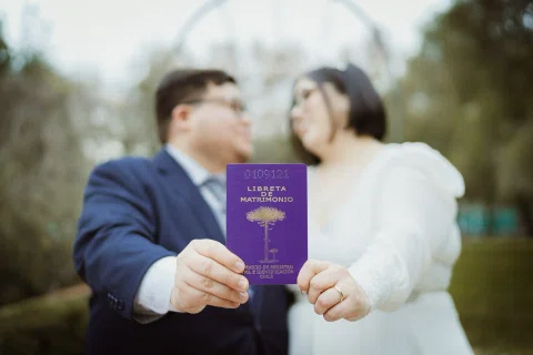 Pequeño cuaderno morado sostenido hacia la cámara, con un hombre y una mujer desenfocados al fondo.