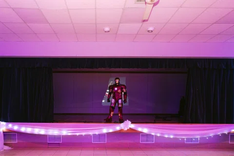 Persona con un disfraz de armadura roja y dorada permanece sola sobre un escenario iluminado en violeta.