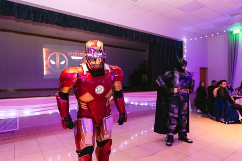 Persona con disfraz de armadura roja y dorada aparece en primer plano mientras otra figura con traje negro permanece al fondo.