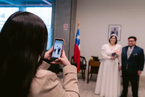 Persona fotografía con un celular a una mujer con vestido blanco y un hombre con traje oscuro.