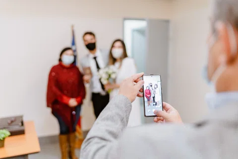 Persona fotografía con un teléfono a un hombre con traje claro y una mujer con vestido blanco que posan al fondo.