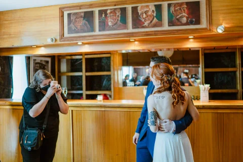 Una persona toma una fotografía de un hombre con traje azul y una mujer con vestido blanco frente a una barra de madera.