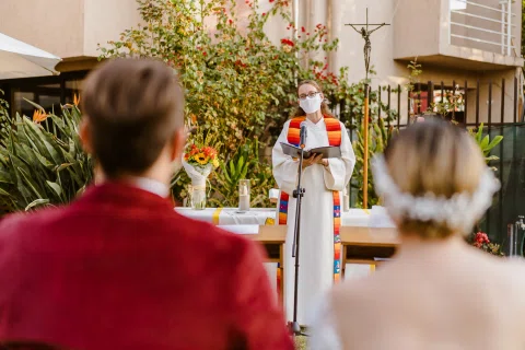 Persona con túnica blanca y una banda de colores permanece frente a un micrófono, con un hombre de chaqueta roja y una mujer de vestido blanco sentados de espaldas.