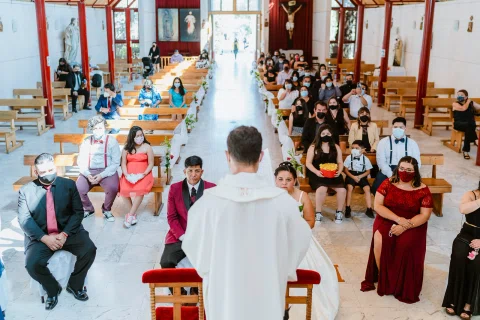 Persona con vestimenta blanca vista de espaldas frente a varias personas sentadas dentro de una iglesia.