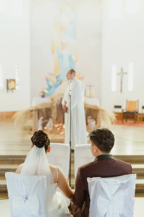 Persona con vestimenta blanca está de pie frente a un hombre y una mujer sentados en una iglesia.