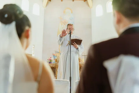 Persona con vestimenta blanca está de pie al frente de un hombre y una mujer sentados dentro de una iglesia.