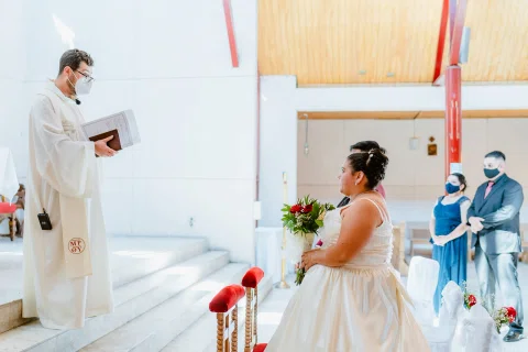 Persona con vestimenta blanca sostiene un libro frente a una mujer con vestido blanco sentada.
