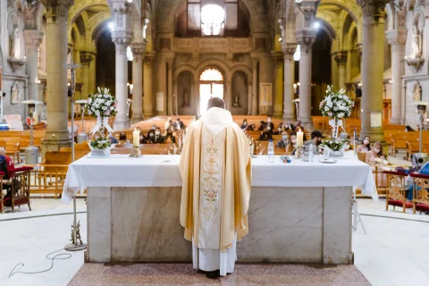 Persona con vestimenta religiosa aparece de espaldas frente a un altar decorado con flores.