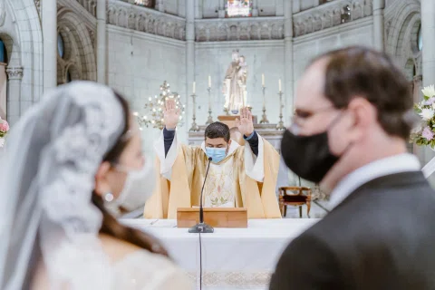 Persona con vestimenta religiosa mantiene ambas manos alzadas, con una mujer con velo y un hombre con traje oscuro desenfocados en primer plano.