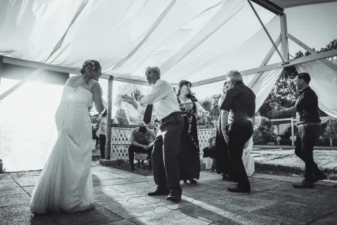 Varias personas bailan bajo una carpa en una fotografía en blanco y negro; una mujer con vestido blanco está entre ellas.