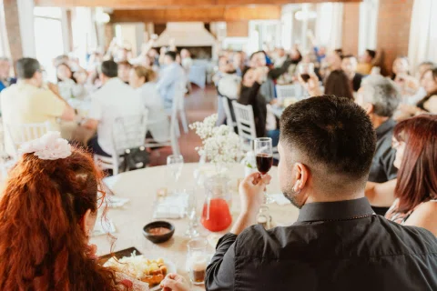 Personas comparten una comida alrededor de mesas dentro de un salón de ladrillo y madera.