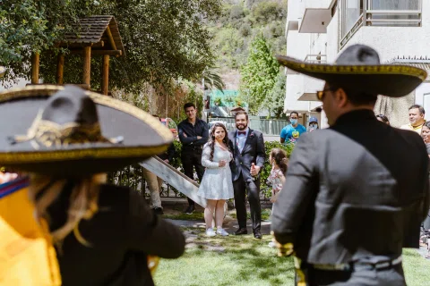 Dos personas con sombreros e instrumentos aparecen de espaldas frente a un grupo reunido en un jardín.