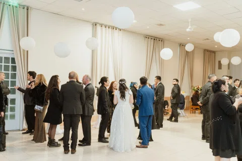 Varias personas conversan de pie en un salón; una mujer con vestido blanco y un hombre con traje azul aparecen cerca del centro.