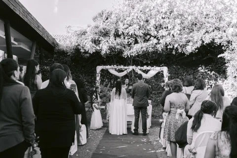 Personas de pie y sentadas observan un arco floral al aire libre, en una fotografía en blanco y negro.